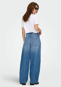 Jeans denimi ușor albastri cu picior larg, talie înaltă, design cu cinci buzunare și o textură ușor decolorată. Poartă un tricou alb și ochelari de soare negri.