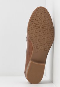 Semelle de chaussure en cuir marron avec motif de marque "Tamaris" répété et taille 37, avec un talon texturé pour une bonne adhérence, inclinée contre un coin blanc.