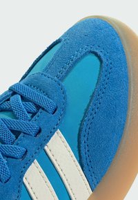 Blue athletic shoe featuring suede and fabric surfaces, a textured white stripe, and a rubber sole. Laces are secured in a geometric pattern.