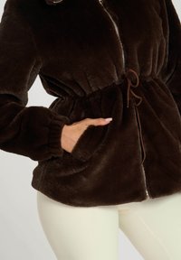 Bruine nepbontjas met een tailleband, ritsluiting en voorzakken. Kenmerkt zich door een zachte textuur en elastische manchetten, gecombineerd met lichtgekleurde broeken.