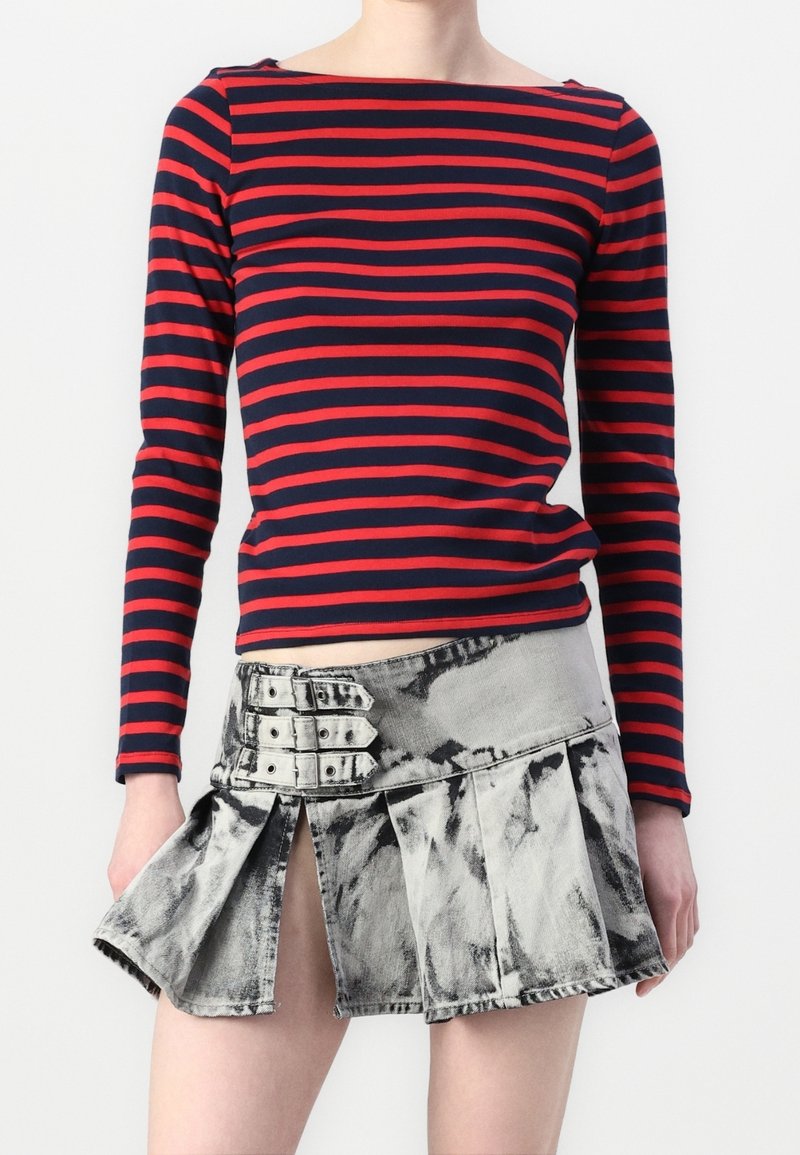 Woman wearing a red and navy striped long-sleeve top and a gray acid-wash pleated mini skirt with three silver buckle straps.