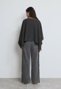 Mujer de espaldas que lleva un poncho gris oscuro con mangas anchas y vaqueros grises holgados con detalles de flecos a los lados.