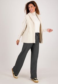 Crème-kleurige fleece vest met een kraag, gedragen over een licht shirt en donkere, losjes zittende broeken. Zwart en geel gestreepte sneakers.