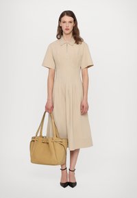 Beige mekko, jossa on lyhyet hihat, vetoketjukaulus ja laskostettu muotoilu, paritettuna keltaisen nahkatote-bagin ja mustien kärki-korkokenkien kanssa.