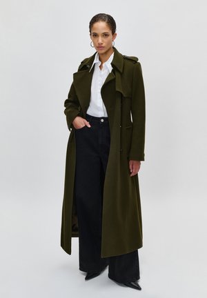Femme portant un long trench-coat vert foncé sur une chemise blanche et un pantalon noir à jambes larges, debout devant un fond clair uni.