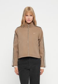 Mulher loira a usar uma jaqueta cropped bege com fecho de correr, com riscas nas mangas em preto e branco e calças pretas, de pé diante de um fundo branco.