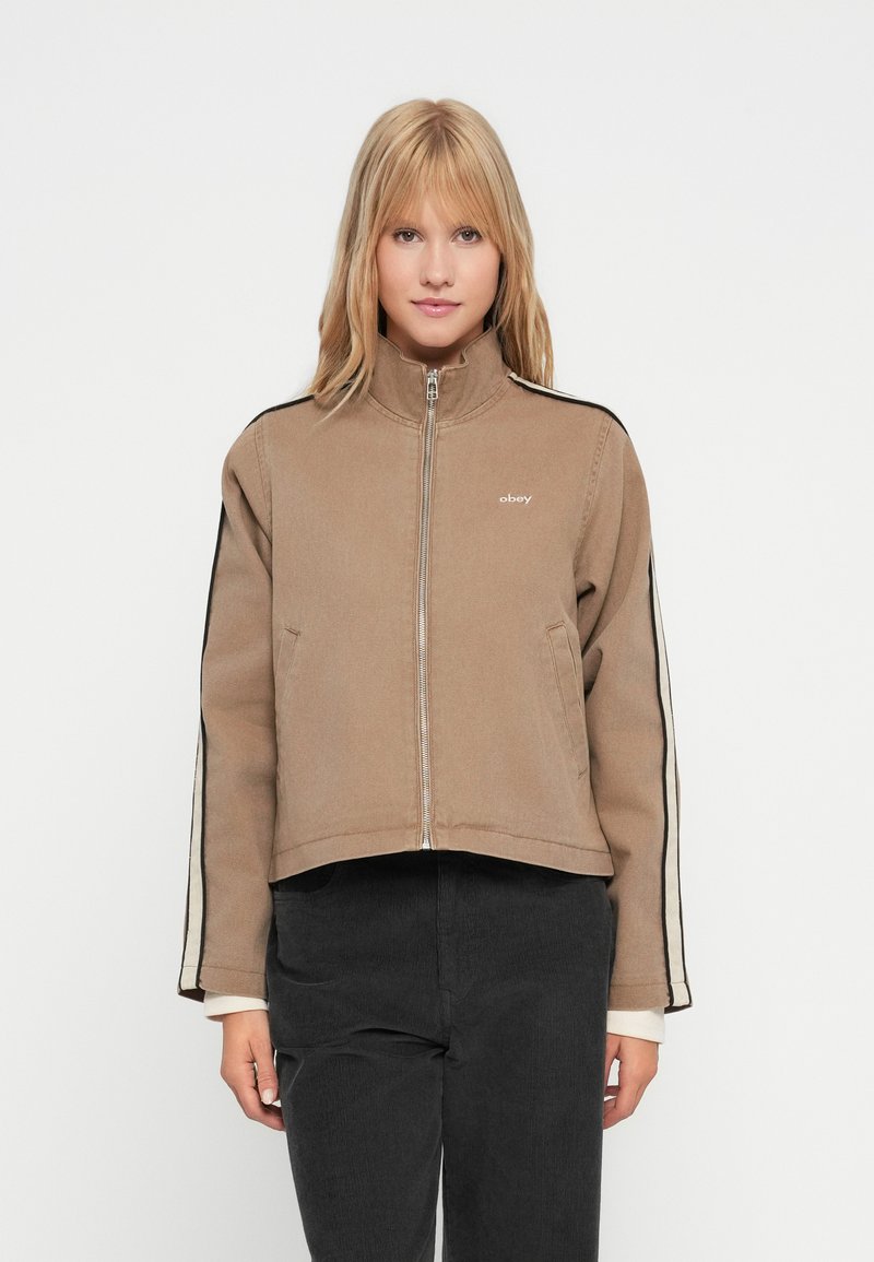 Mulher loira a usar uma jaqueta cropped bege com fecho de correr, com riscas nas mangas em preto e branco e calças pretas, de pé diante de um fundo branco.