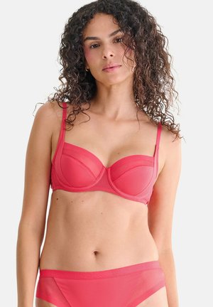 Femme aux cheveux bouclés portant un ensemble assorti de soutien-gorge et culotte rouges, debout devant un fond clair uni.