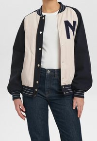Veste universitaire avec corps crème et manches bleu marine, dotée de poignets côtelés rayés, de boutons-pression et d'un grand "N" brodé sur la poitrine.