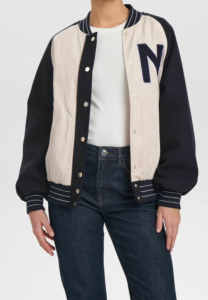 Veste universitaire avec corps crème et manches bleu marine, dotée de poignets côtelés rayés, de boutons-pression et d'un grand "N" brodé sur la poitrine.