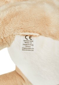 Beige plush toy with soft, textured fur. A care label attached shows CE marking and brand details. White underbelly contrasts with main color.