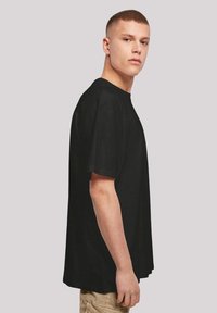 Schwarzes Baumwoll-T-Shirt mit klassischem Schnitt, kurzen Ärmeln und rundem Halsausschnitt, von der Seite an einem Modell vor einem neutralen Hintergrund präsentiert.