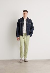 Jeansjacke mit Reißverschlusstasche, weißes T-Shirt und hellgrüne Jogginghose. Model in grauen Turnschuhen steht vor einem schlichten Hintergrund.