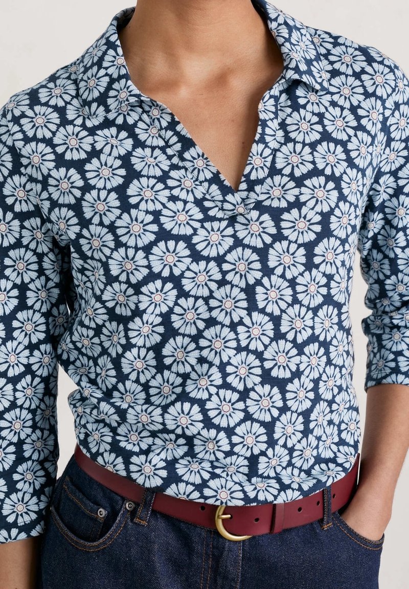 Chemise marine avec motif floral blanc, design col, manches trois-quarts et une ceinture bordeaux avec des accessoires dorés. Texture en mélange coton.