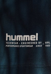 Hummel-logo og tekst på mørkt stof med teksten "Techwear - udviklet af HML, performance sportswear siden 1923."