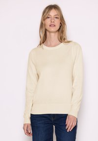 Maglione color crema realizzato in morbido tessuto, con scollo rotondo, maniche lunghe e polsini e orlo a costine. Indossato con jeans blu scuro.