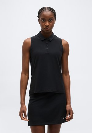 Ženska z vlečenimi lasmi, oblečena v črno brezrokavnico z ovratnikom in črno krilo Nike, stoji pred enostavnim svetlim ozadjem.