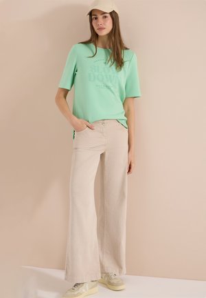 Giovane donna con cappello beige, maglietta verde menta con scritta, pantaloni beige larghi e sneakers beige, in piedi contro un muro semplice.