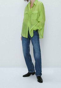 Lime green, sheer button-up shirt with two chest pockets, paired with blue straight-leg jeans and black pointed-toe boots.