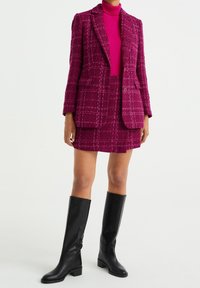 Completo giacca e gonna mini in tweed fucsia con un motivo a griglia, abbinato a stivali al ginocchio in pelle nera. Texture liscia, vestibilità su misura.