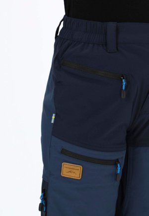 Marineblå cargobukser med elastisk taljebånd, flere lynlåslommer og en tanfarvet logo-patch. Materialet virker let og slidstærkt.