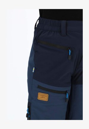 Marineblaue Cargo-Hose mit elastischem Bund, mehreren Reißverschlusstaschen und einem braunen Logo-Patch. Das Material wirkt leicht und strapazierfähig.