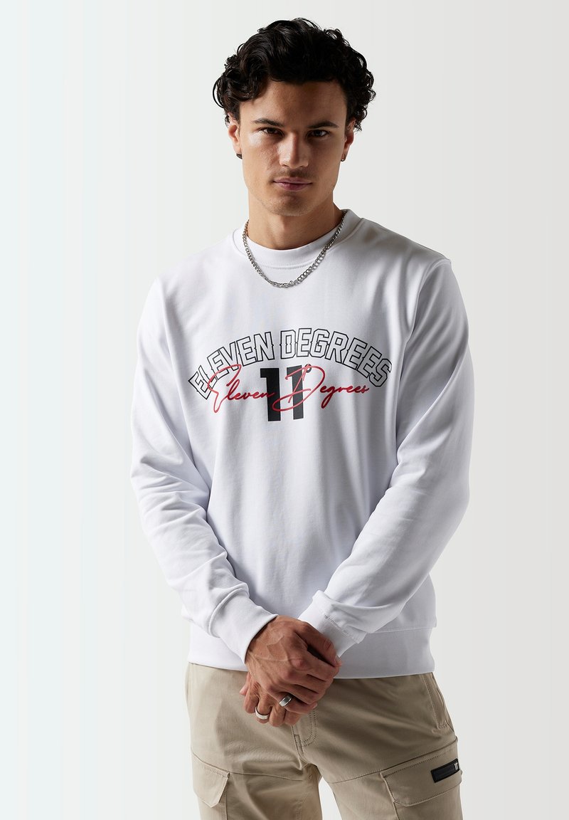 Sudadera de cuello redondo blanca con un audaz gráfico en negro y rojo de "Eleven Degrees", mangas largas y puños acanalados. Modelo posando con las manos entrelazadas.