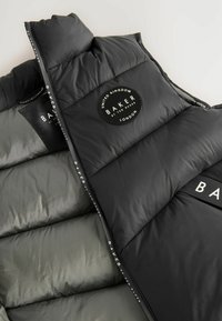 Crna puffer prsluk sa siva obloga, sa okruglim logotipom sa natpisom "BAKER by TED BAKER." Kvadrični dizajn s izraženim sekcijama.