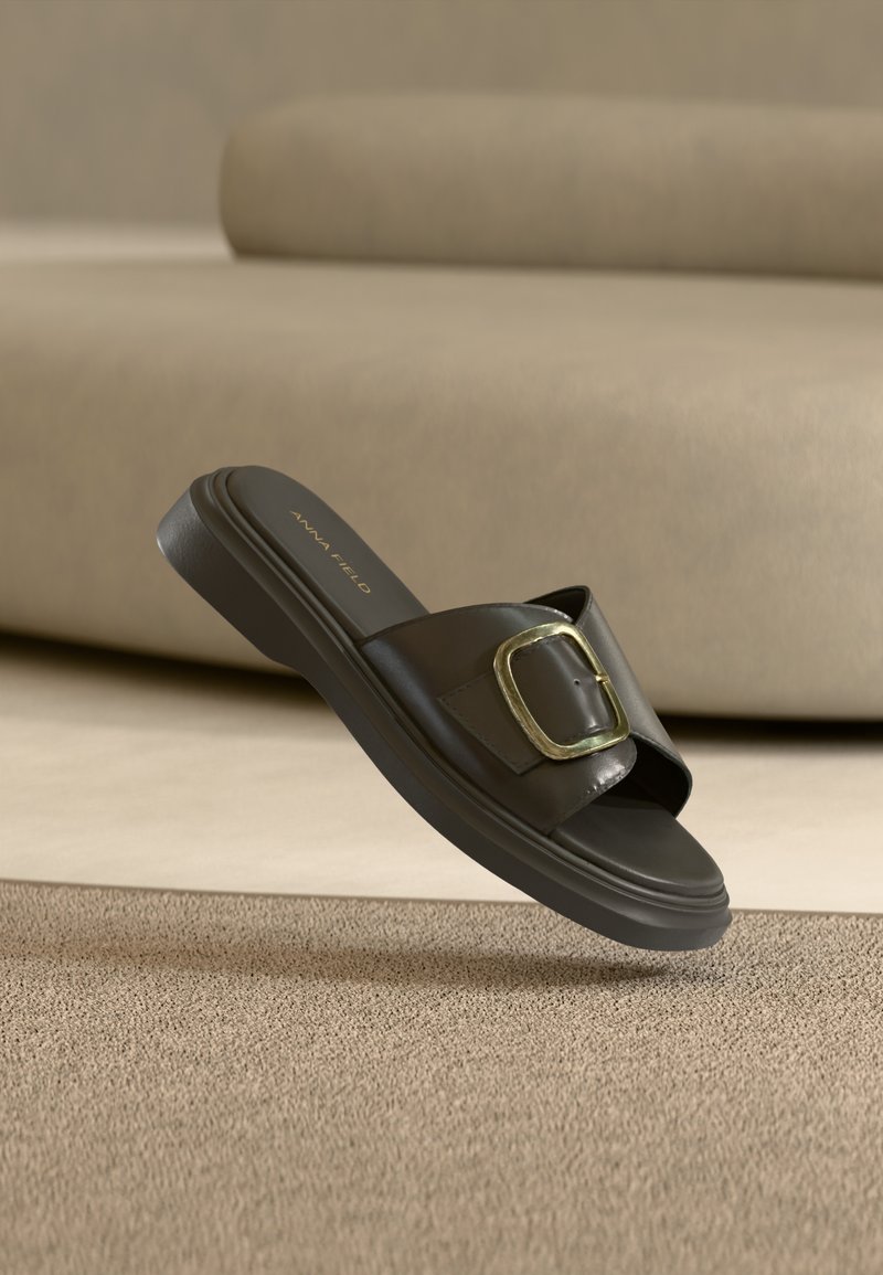 Μαύρη σανδάλι χωρίς λουράκι με φαρδιά λωρίδα και μεγάλο χρυσό πόμολο, ακουμπισμένο σε μπεζ μοκέτα κοντά σε καφέ καναπέ.