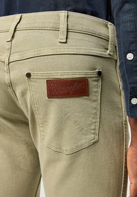 Los pantalones vaqueros Wrangler en color caqui cuentan con una tela texturizada, una etiqueta de cuero marrón en contraste y un diseño de bolsillo distintivo con un patrón en zigzag.