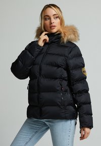 Svart pufferjakke med hette av faux pels, glidelås foran, sidelommer og quiltet design. Utsmykket med en sirkulær logo på ermet.