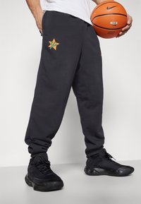Šedé tepláky s oranžovou hvězdnou grafikou a zeleným detailem na levé kapse, v kombinaci s černými sportovními botami. V ruce drží basketbal.