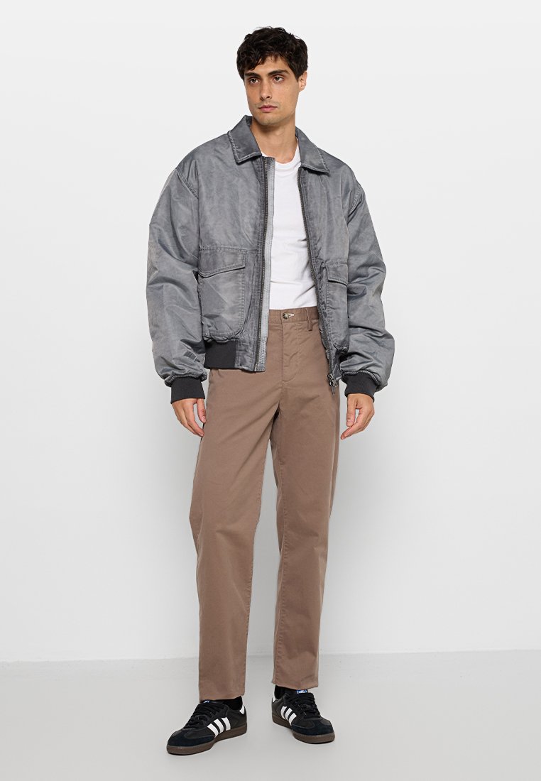 Graue Bomberjacke mit Reißverschluss, zwei Fronttaschen und ribbed Manschetten, getragen über einem weißen T-Shirt und hellbraunen Hosen, mit schwarzen Sneakers.