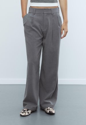 Pantaloni grigi a gamba larga con pieghe frontali, passanti per cintura e tasche laterali. Abbinati a scarpe basse con fantasia. Texture liscia e vestibilità sartoriale.