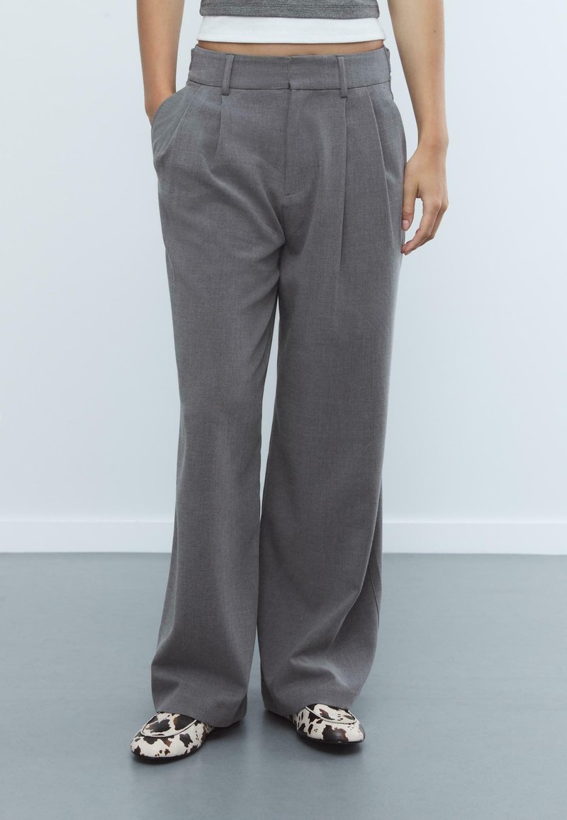 Graue weit geschnittene Hose mit vorderen Falten, Gürtelschlaufen und seitlichen Taschen. Kombiniert mit gemusterten flachen Schuhen. Glatte Textur und taillierte Passform.