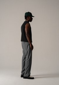 Schwarzes ärmelloses Top, graue Hose und schwarze Schuhe mit weißen Akzenten. Die Person trägt eine grüne Mütze. Einfache, gedeckte Farbpalette.
