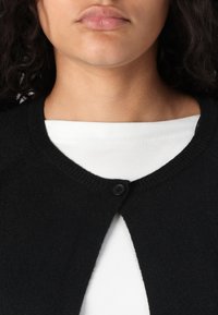 Black, knitted cardigan with a round neckline, featuring a single button closure at the front, worn over a white shirt.