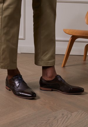 Chaussures habillées en cuir marron foncé portées avec un pantalon kaki court sur un sol en bois clair, avec une chaise en bois partiellement visible en arrière-plan.