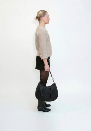 Femme debout de profil, portant un pull beige, une jupe noire, des collants, des bottes, tenant un grand sac à main noir avec une anse tressée sur fond blanc.
