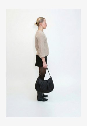 Femme debout de profil, portant un pull beige, une jupe noire, des collants, des bottes, tenant un grand sac à main noir avec une anse tressée sur fond blanc.