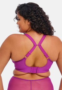 Sujeción morado con un diseño deportivo que presenta tirantes anchos, herrajes ajustables y un cierre de gancho y ojal en la parte posterior. Textura suave.