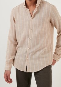 Béžová košile s dlouhým rukávem na knoflíky s bílými vertikálními pruhy, vyrobená z texturované látky, s klasickým límcem a zapínáním na knoflíky vpředu.