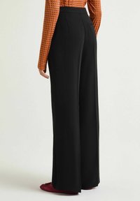 Pantalon noir à jambes larges avec une texture lisse et une taille haute. Coutures détaillées le long des coutures et une poche arrière. Stylé avec une chemise à motifs.