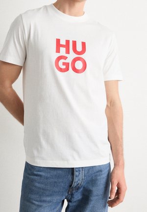 Mann trägt ein weißes kurzärmeliges T-Shirt mit fettgedrucktem roten „HUGO“-Schriftzug und blaue Jeans, steht vor einem schlichten hellen Hintergrund.