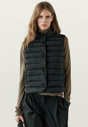 Ung kvinde med langt hår iført en sort quiltet vest over en olivengrøn langærmet skjorte og sorte bukser, der holder en lille sort taske.