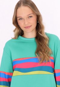 Hellgrüner Sweatshirt mit Rundhalsausschnitt und langen Ärmeln. Verfügt über wellenförmige Streifen in Rot, Blau und Gelb über die Brust.