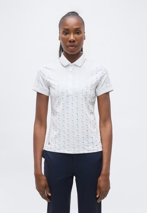 Woman wearing a white short-sleeve polo shirt with small gold palm tree patterns and dark blue pants, standing against a plain white background.