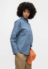 Γυναίκα με κατσαρά μαλλιά φοράει μπλε ριγέ πουκάμισο με κουμπιά και μπεζ παντελόνι, κρατώντας μια πορτοκαλί κάπα στο δεξί της χέρι.