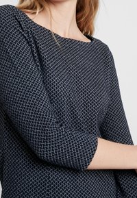 Laivastonsininen toppi, jossa on geometrinen kuvio, kolmanneksi viimeiset hihat ja leveä kaula-aukko. Teksturoitu kangas, jossa on sileä pinta.