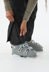 Grijze skibotten met plastic gespen en een gestructureerd ontwerp, gecombineerd met zwarte waterdichte broeken die worden aangepast. Gladde, taps toelopende pasvorm zichtbaar.